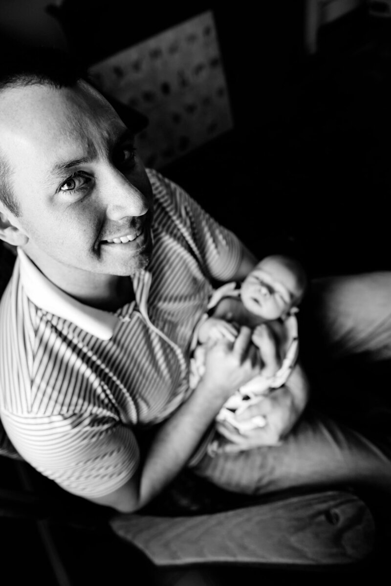 A black and white photo from a high angle capturing a man looking up and smiling as he holds a newborn baby in his arms.
