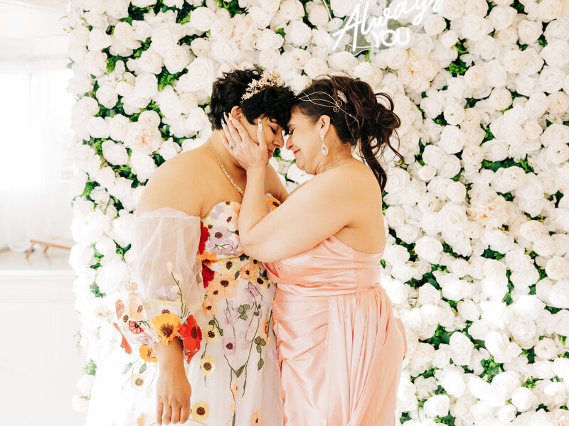 Two women stand close together in front of a white flower wall, gently touching foreheads and smiling. One wears a floral off-shoulder dress, the other a pink gown. Captured by a Colorado Wedding Photographer, the moment feels joyful and intimate.