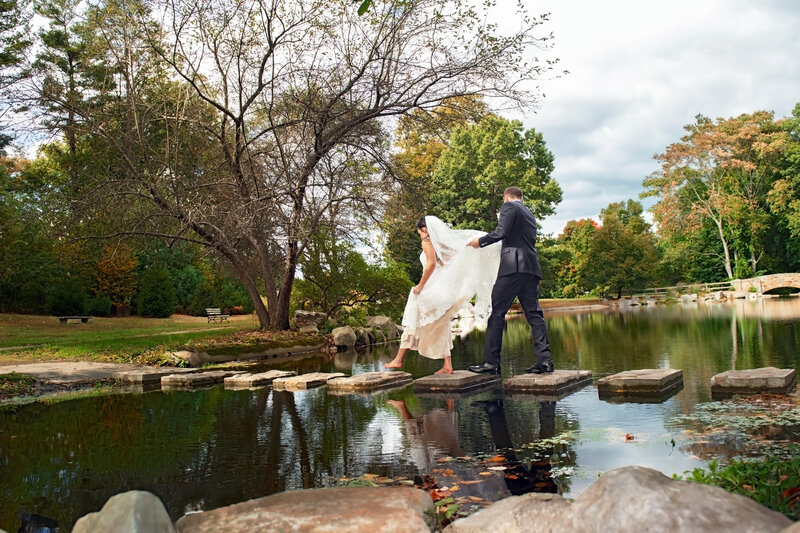 A groom holding a brides dress up as they walk over a river