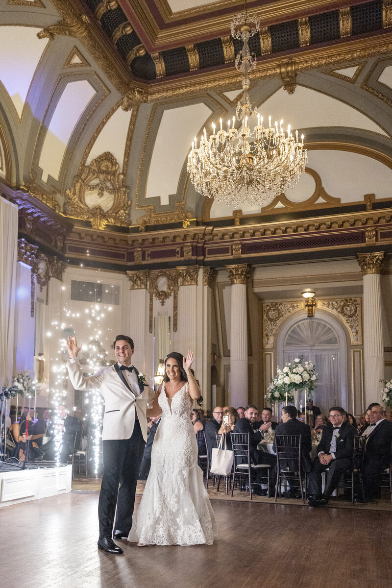 A bride and groom wave and smile during their wedding reception in an ornate ballroom. The room features a grand chandelier, elegant architectural details, and seated guests watching them.