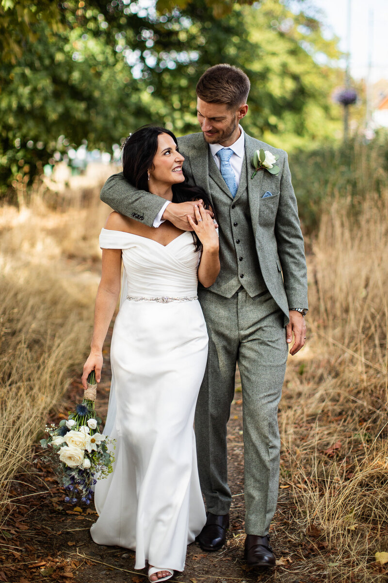 Tall groom wrapping arm around bride in a field of dry grasses