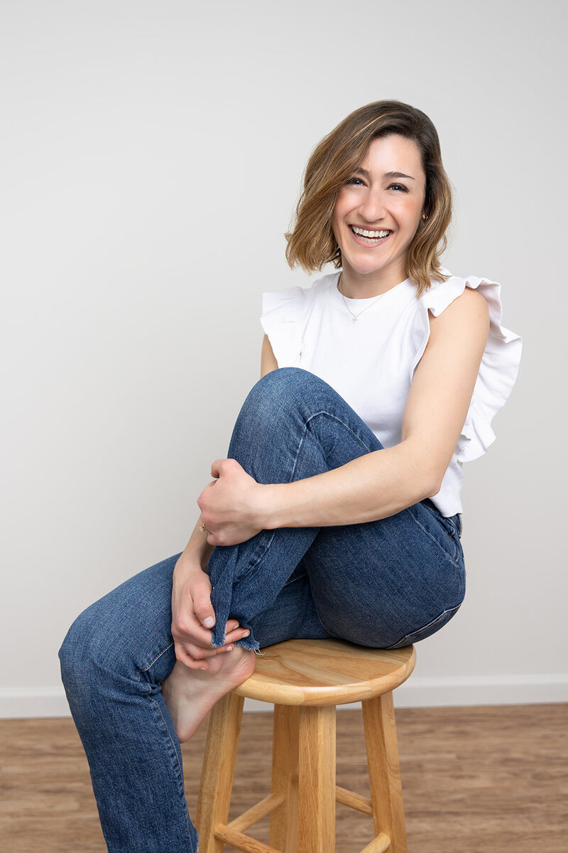Photo of the owner, Gabrielle, she has shoulder length hair, and is wearing a white top and jeans, and sitting on a stool and smilling at the camera