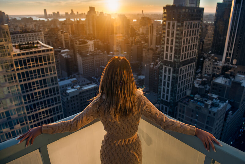 Young woman standing on a balcony enjoying the sun setting over the cityscape.