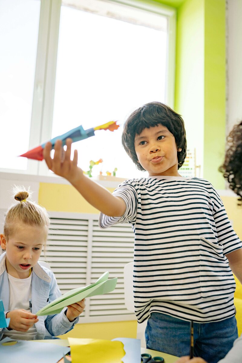 A young boy in a striped shirt enthusiastically holds up a handmade paper rocket, while another child looks curiously at their own paper craft.