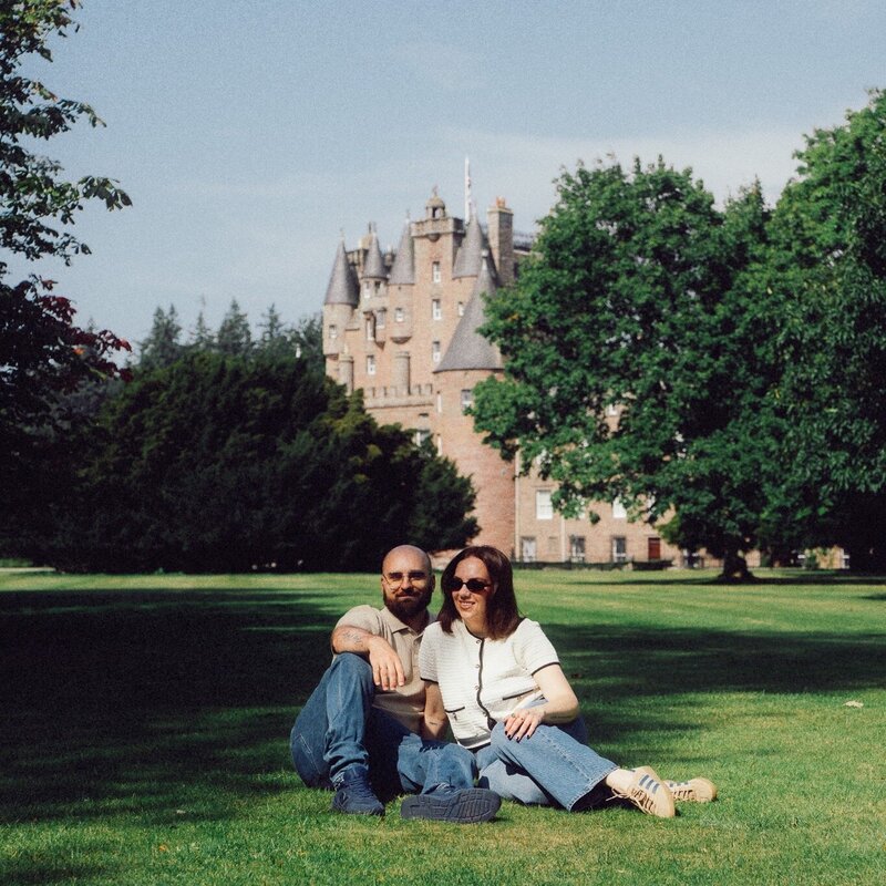 A husband and wife photography duo sit together on the grass in front of a historic castle, relaxed and smiling in the soft daylight.