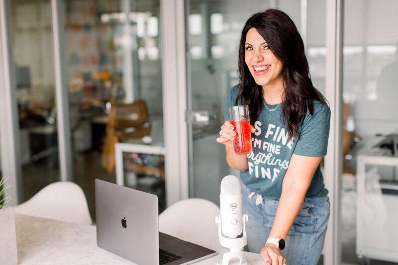 woman with short dark haor wearing a yellow floral shirt and bright lipstick laughing with iced coffee and blue yeti microphone in front of her