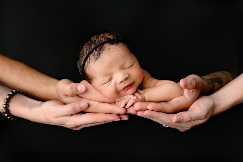 newborn girl wearing a black bow is  laying in parents hands on black background