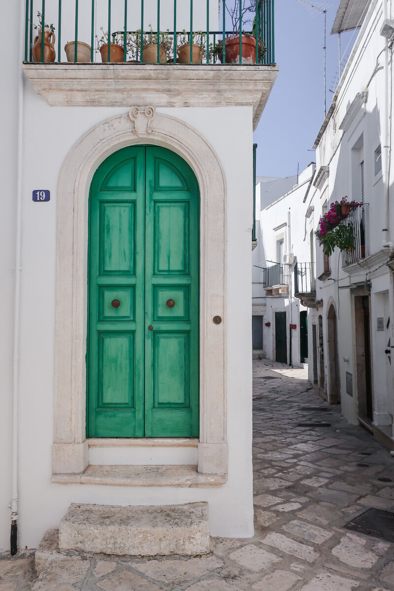 A green door sits under a high arch off of a narrow cobblestone alley of white residences