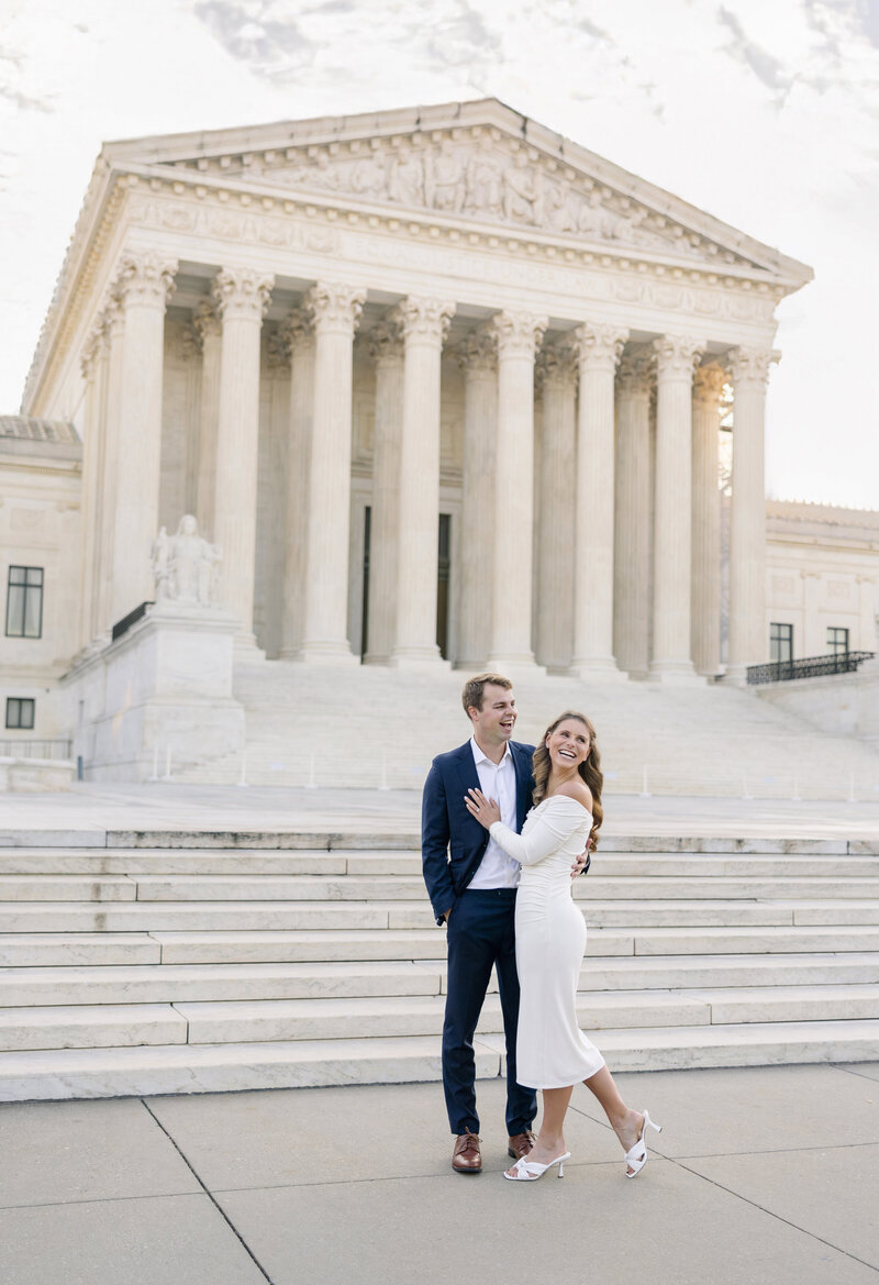 A couple poses in front of The Supreme Court, a large neoclassical building with tall columns. The woman is wearing a white dress and heels, while the man is in a navy suit. They are smiling at each other beneath a set of stairs.