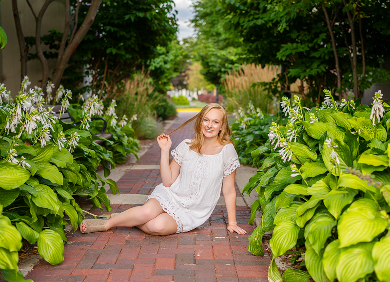 high school senior girl wearing white dress sitting in alleyway with flowers and bushes on each side of her