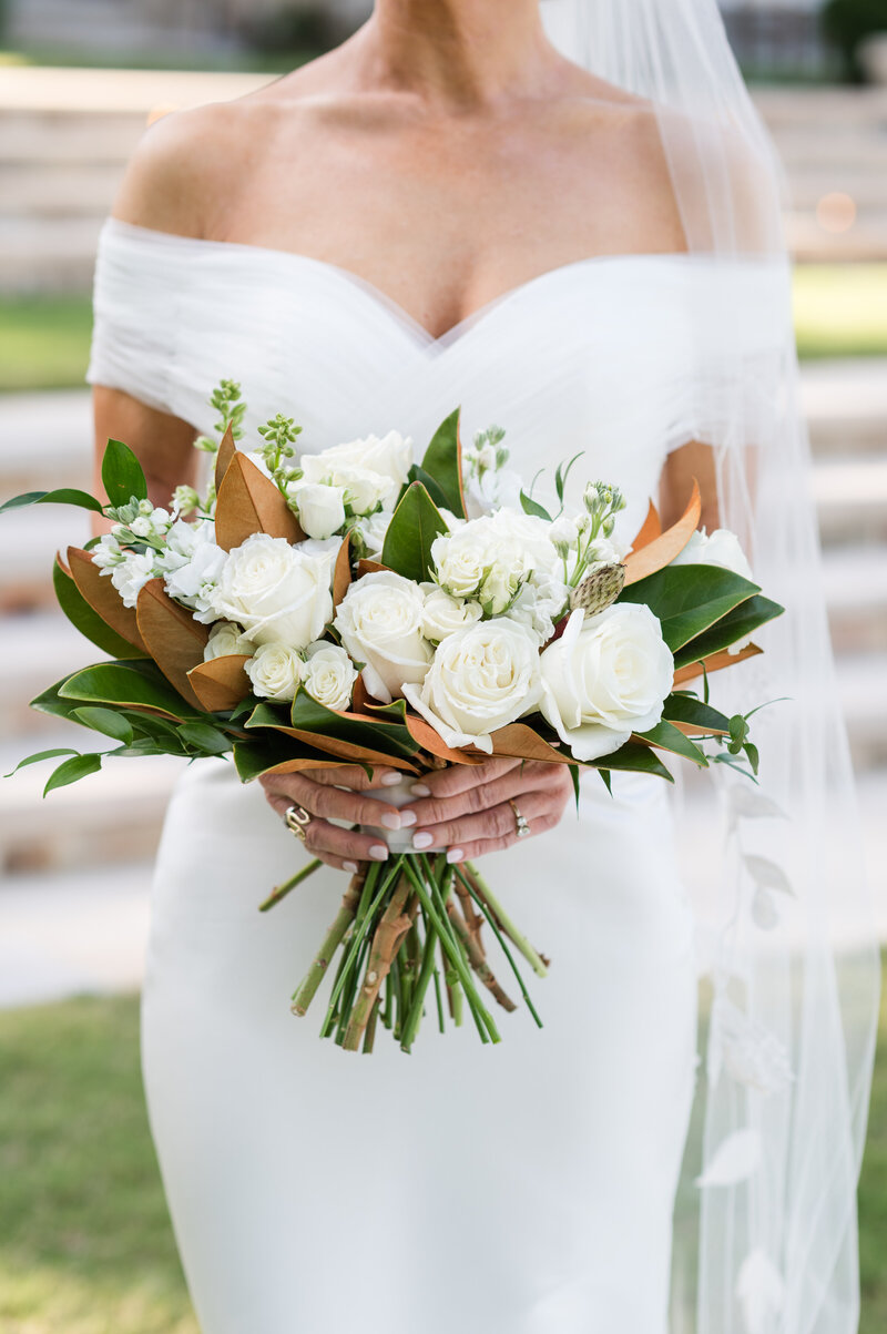 Bride holding wedding bouquet of white roses and green magnolia leaves.