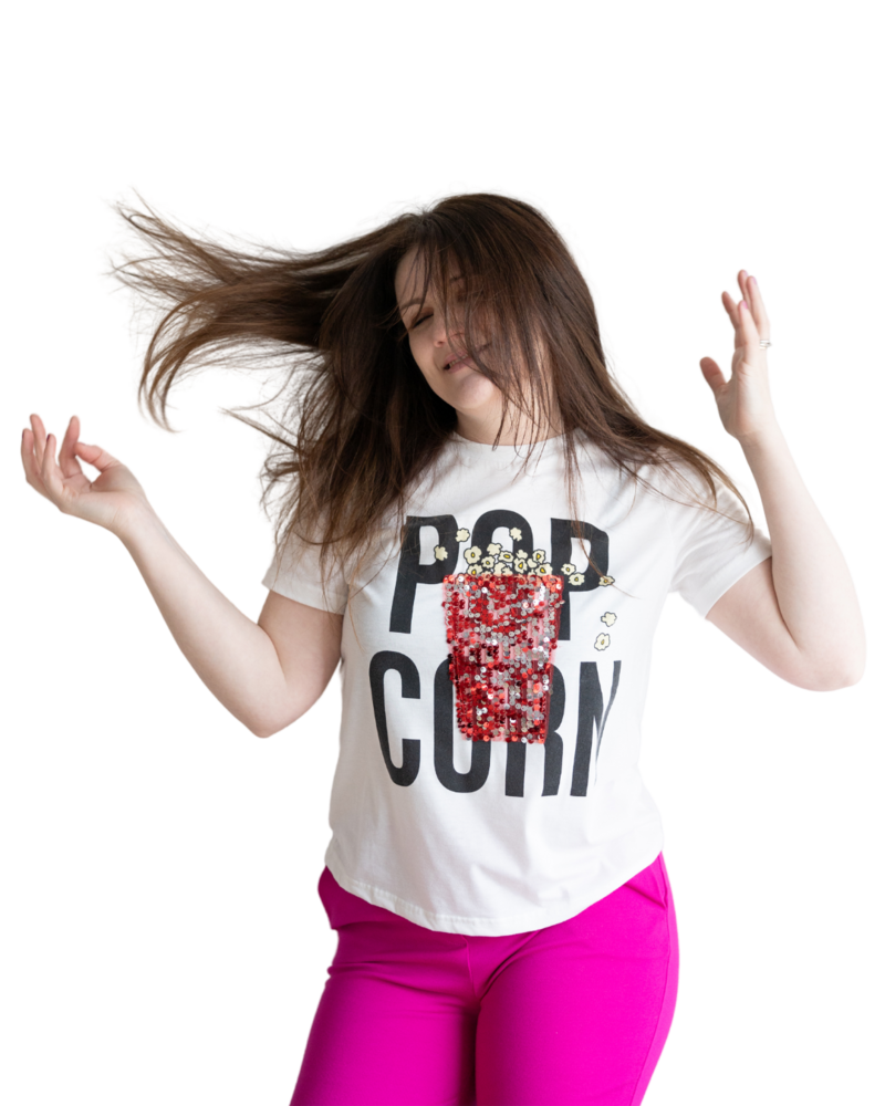 Emily wearing hot pink pants and a shirt that says pop corn with an image of glittery popcorn on it. She is flipping her hair in the air with her eyes closed and arms up in the air