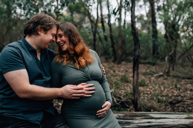 Husband and wife sitting on log laughing together during maternity session in Melbourne bushland.