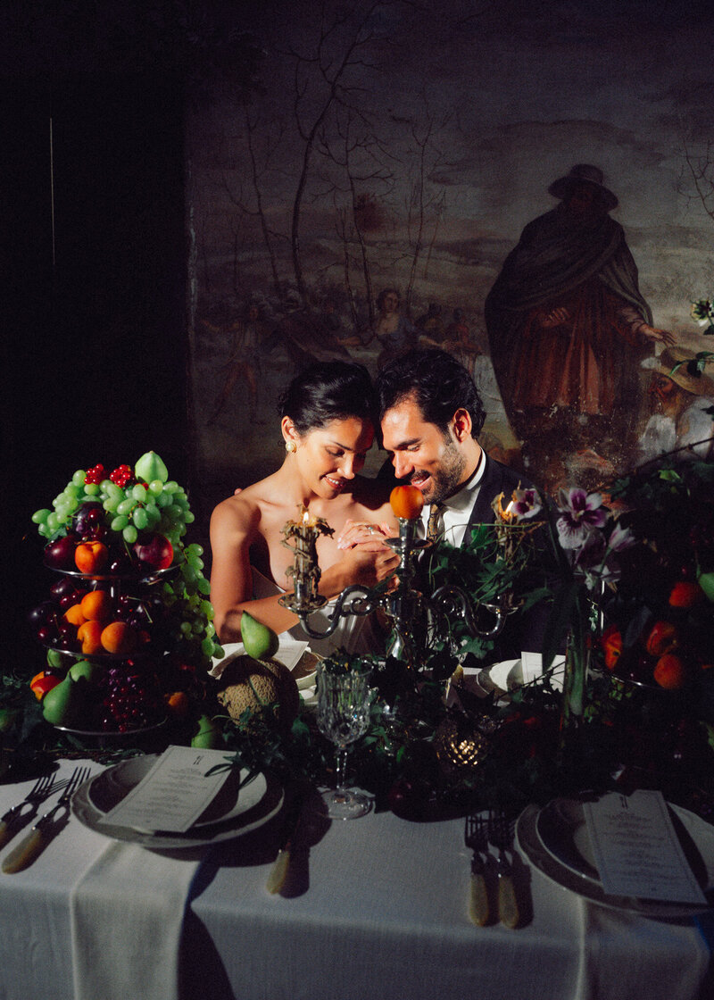 Intimate candlelit wedding moment at an elegant tablescape with abundant fruit and florals, captured in a romantic, cinematic style by a luxury documentary wedding photographer UK.