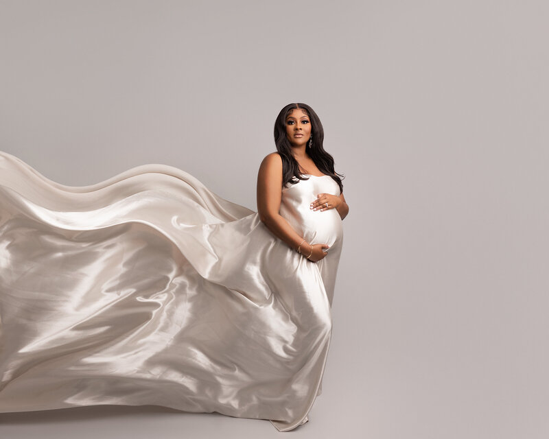 Woman poses for NYC fine art maternity photos. She is draped in a white silky sheet that is cascading and blowing behind her. She is smiling at the camera with one hand atop and one hand below her bump. Captured by best NYC maternity photographer Katie Marshall.