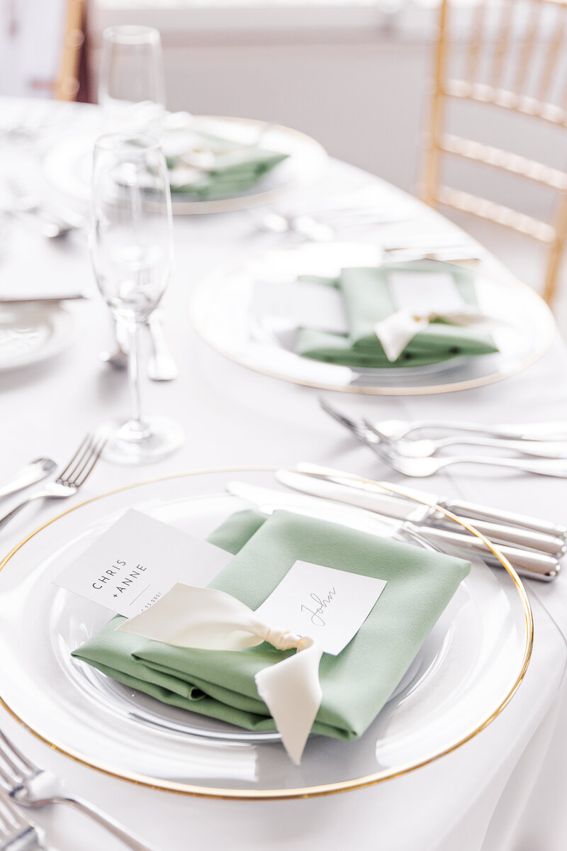 place settings on a round table with white linens and a sage green napkin decorated with a calligraphy name card tied with an off white ribbon