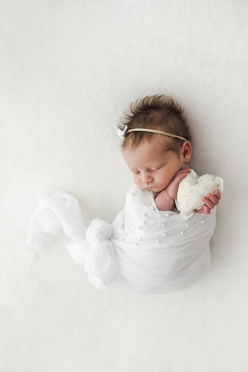 A peaceful newborn wrapped in white, cradling a soft toy, with a tiny bow adorning her delicate hair.