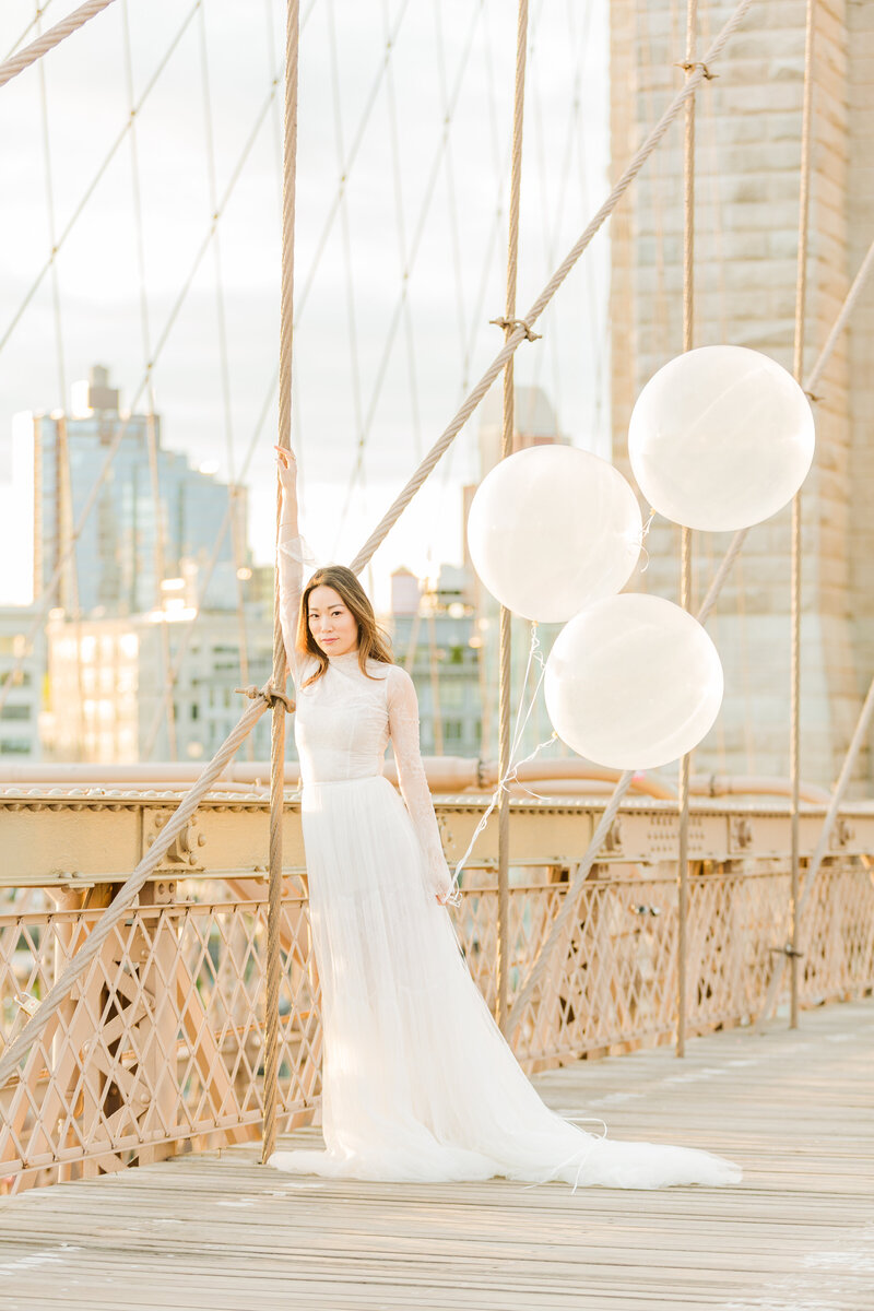 A bride stands on a bridge with her arm resting along the trusses with the New York City skyline in the background. Captured by NYC elopement photographer Lia Rose Weddings