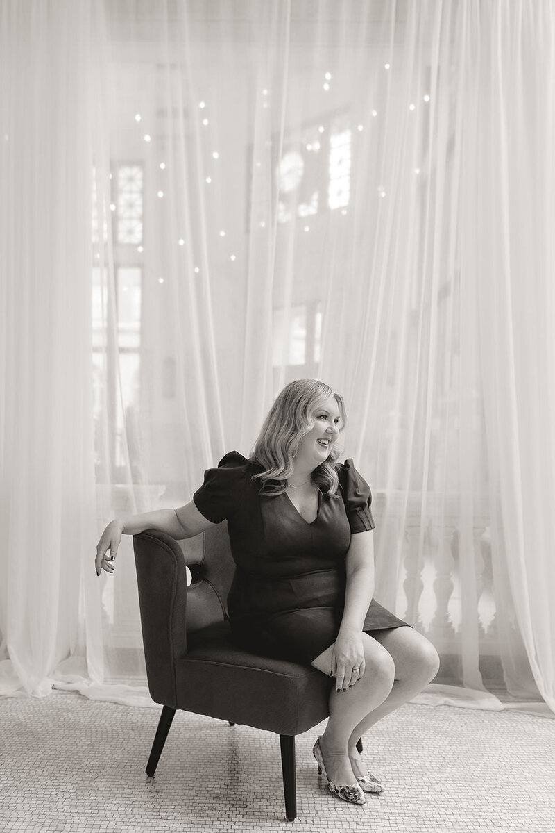 Career coach, Melissa Lawrence, sits in a chair in front of drapes and smiles looking into the distance