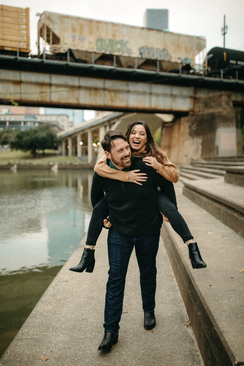 man holding woman on his back standing next to a river with bridge in the background