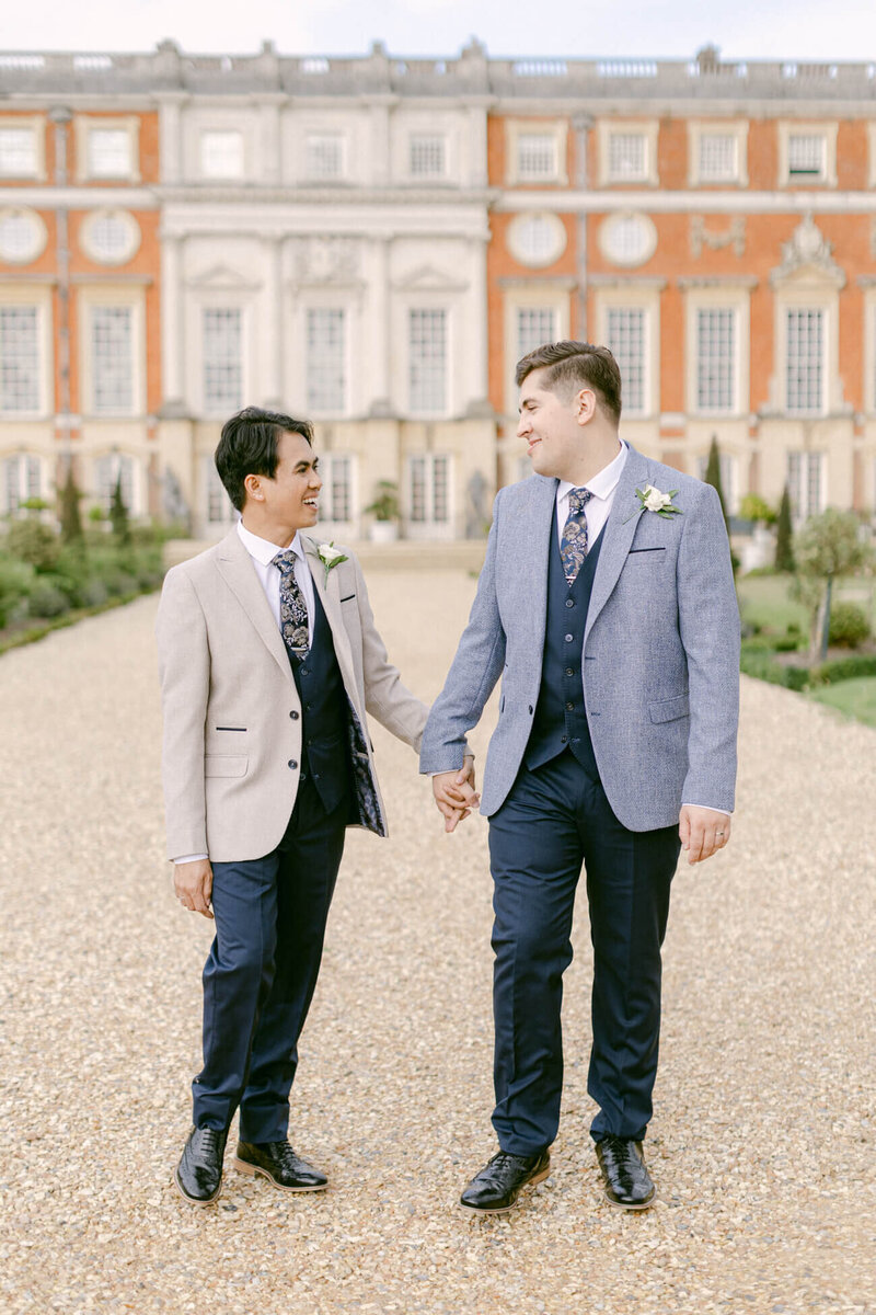 a couple’s portrait at a luxury gay wedding showing two grooms holding hands and walking in the grounds of hampton court palace with the palace behind them