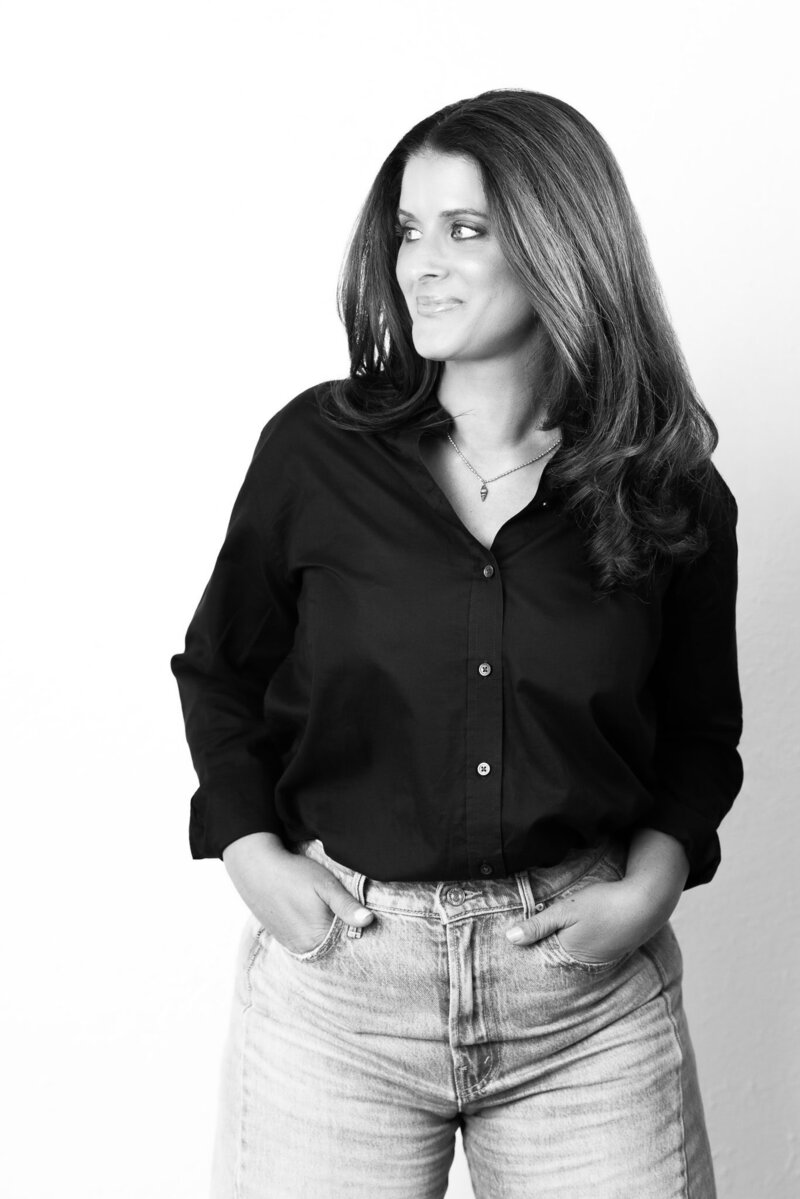 Black & White photo. Woman in black shirt and jeans looking away from camera against white wall.
