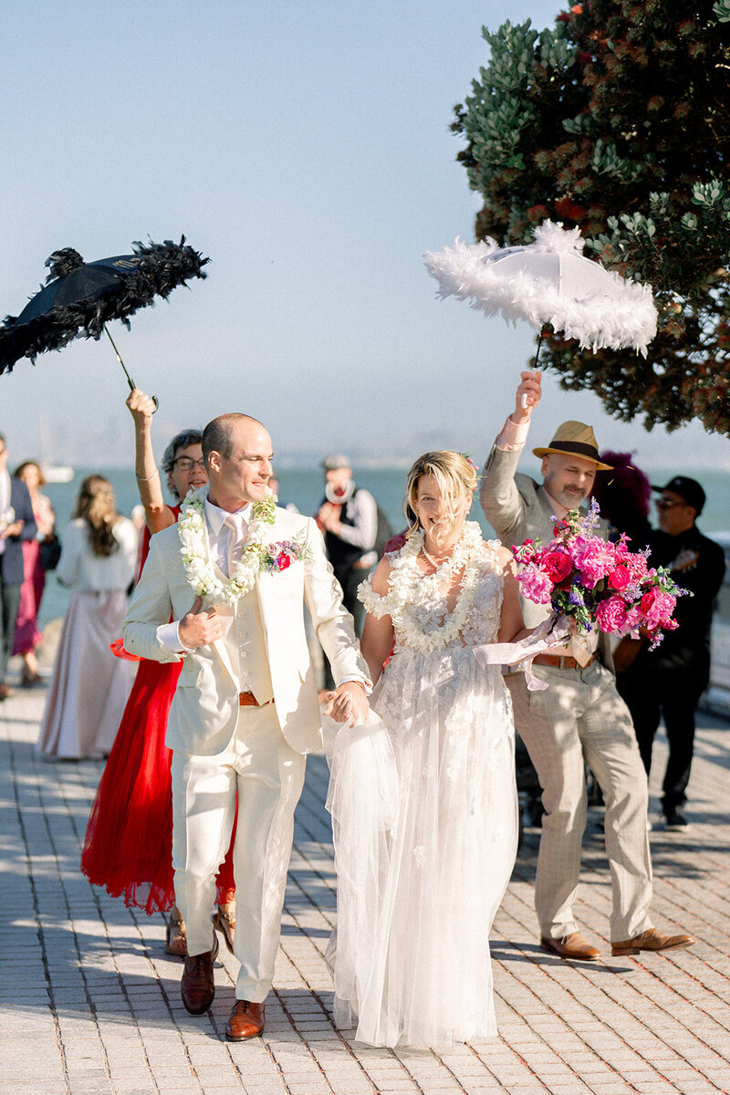 Bride and groom lead a joyful second line parade with parasols at their Bay Area wedding celebration, captured in documentary wedding photography style.