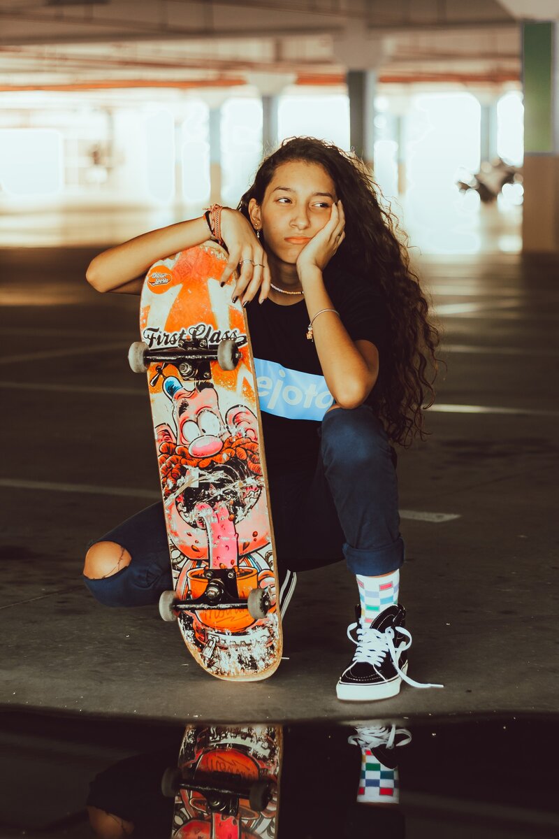 A teen kneels on the pavement in a parking garage, with their right hand resting on the short end of a skateboard. Their chin rests in their left hand, and they have a frustrated expression.
