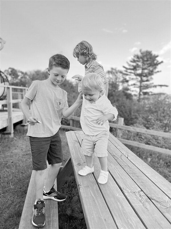 black and white pictures of two older brothers helping their toddler-age brother walk along a picnic table