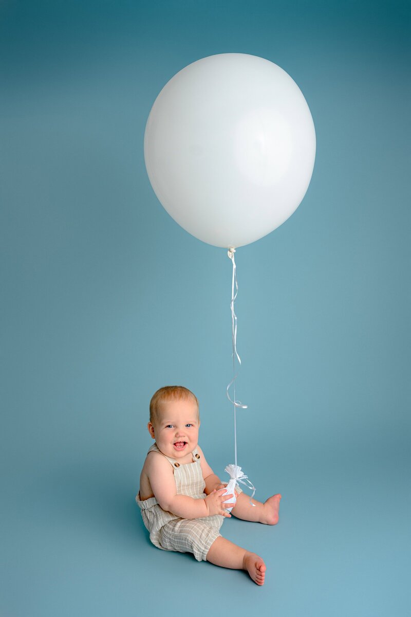 little boy sits in brown overalls holding large white balloon