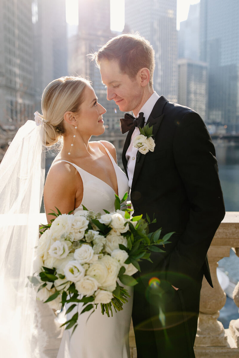 Woman with blonde hair and white rose bouquet looks into groom's eyes on sunny wedding day in Chicago overlooking the skyline.