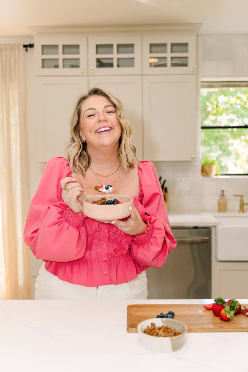 Arch shaped image of Mollie Mason in a bright pink top holding a bowl of food and spoon with fruit on the counter