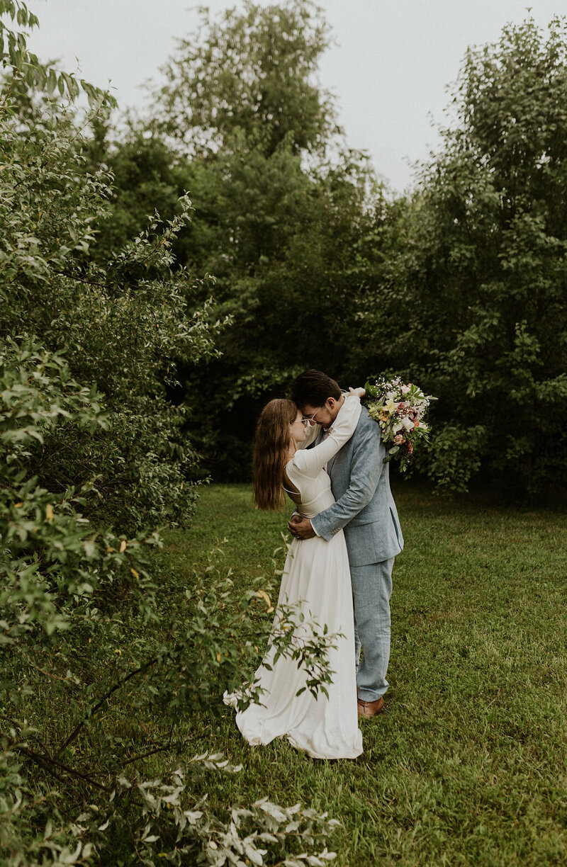 Bride and groom hugging and resting their foreheads together stand in lush green trees
