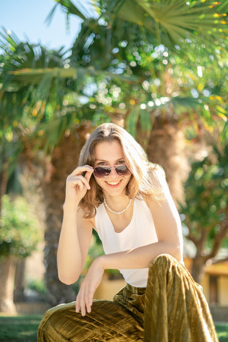High school senior portraits of a girl in a white tank top and gold pants underneath palm trees