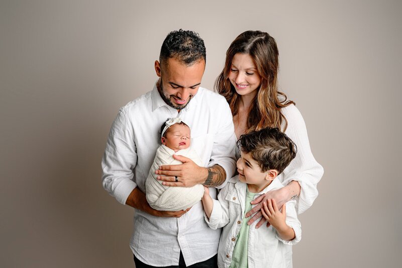 family of 3 standing together looking at thier newborn baby girl wrapped in cream