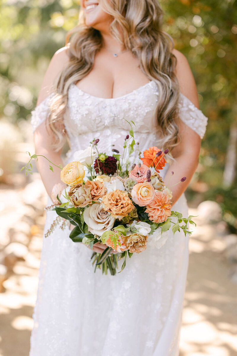 Bride holding a bouquet of warm-toned flowers in an outdoor setting.