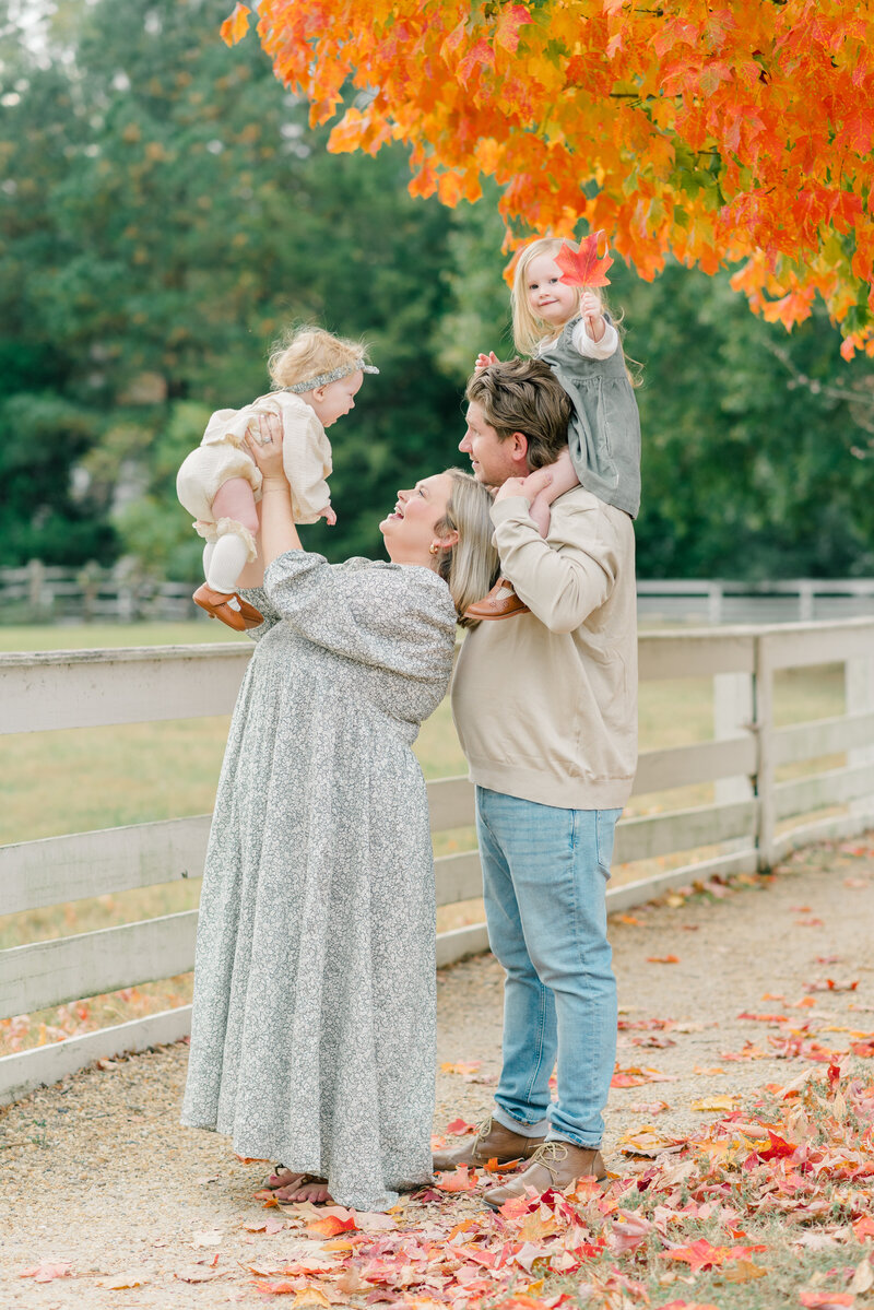 Woman & man hold daughters and smile under fall trees.