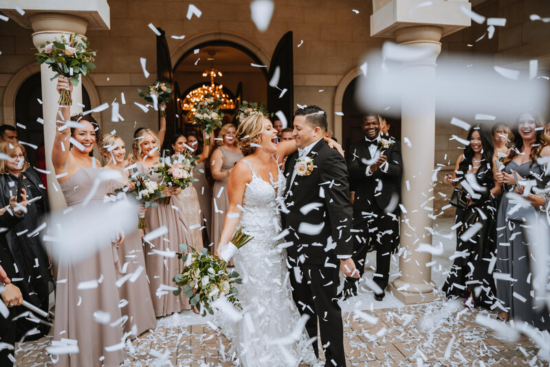 bride and groom smiling at each other surrounded by guests and wedding party with confetti in the air