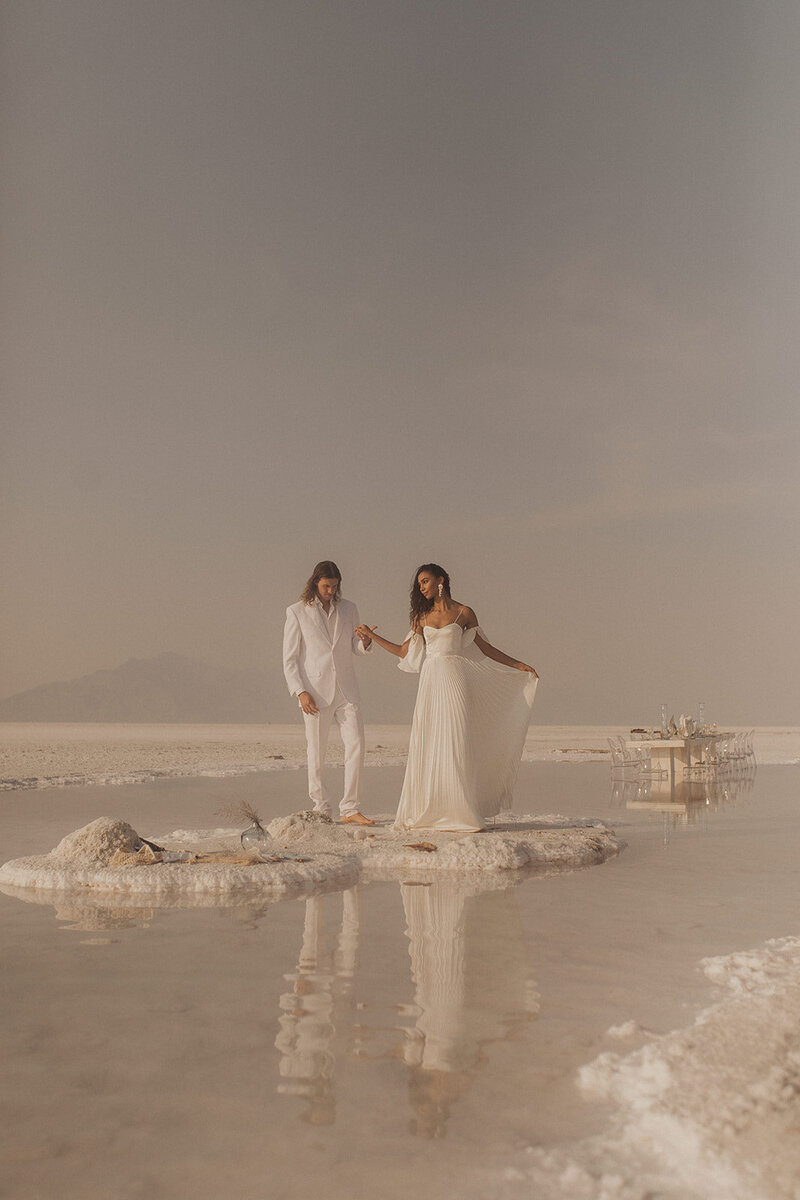 A couple standing apart on a salt flat with a dining table set in the distance.