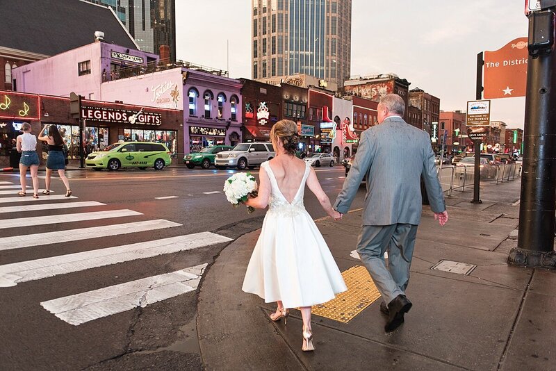 The bride and groom cross Broadway in Nashville hand in hand. They walk away from the camera with the black and white stripes of the crossing lane leading the way to Tootsie's. The bride is wearing a tea length white wedding dress with a plunging back and gold stilettos. She is carrying a large bouquet of white flowers. The groom is wearing a light gray suit.  The Nashville Skyline is in the background.