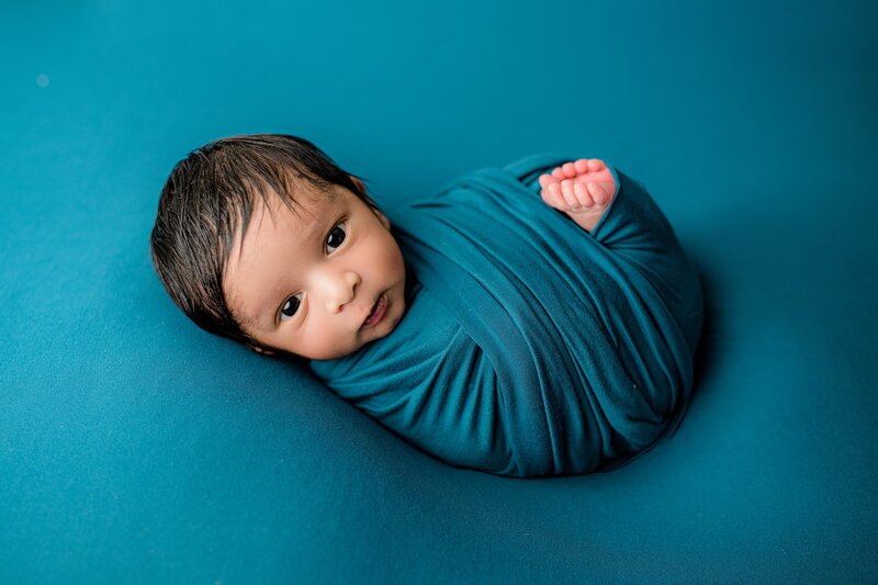 Newborn baby boy is wrapped in a teal wrap with his eyes wide open posed on a teal background