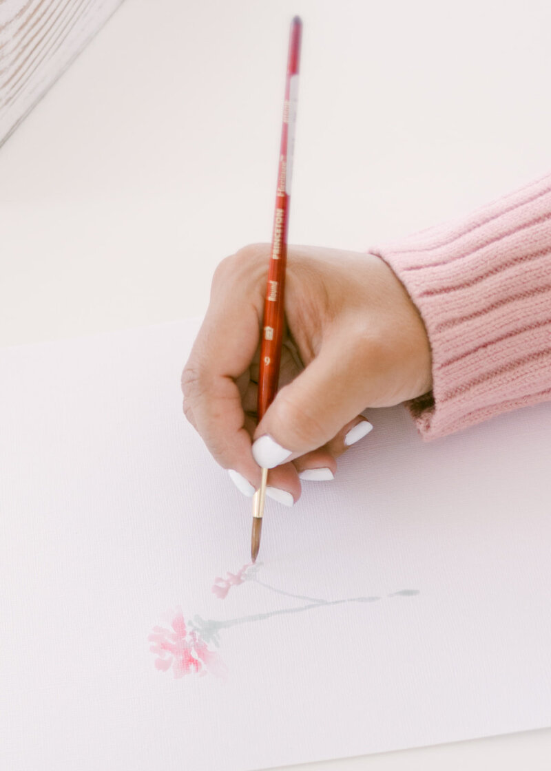 A hand holding a watercolor brush delicately painting a pink flower