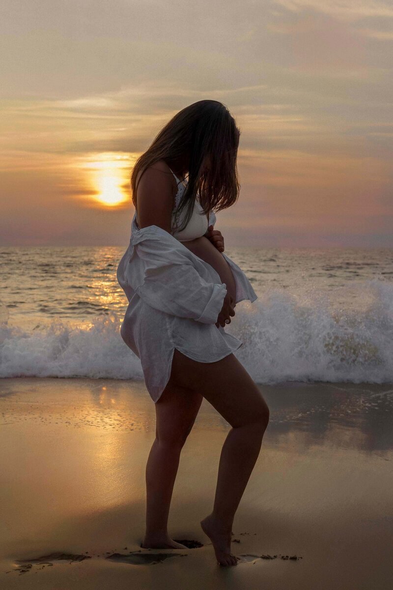 femme enceinte de profil en chemise blanche sur la plage avec un coucher de soleil