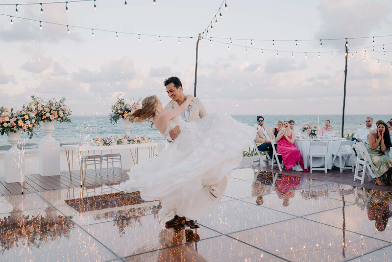 A jubilant groom lifts his bride in a joyous dance at their beach wedding reception, surrounded by cheering guests and a romantic, glittering atmosphere.