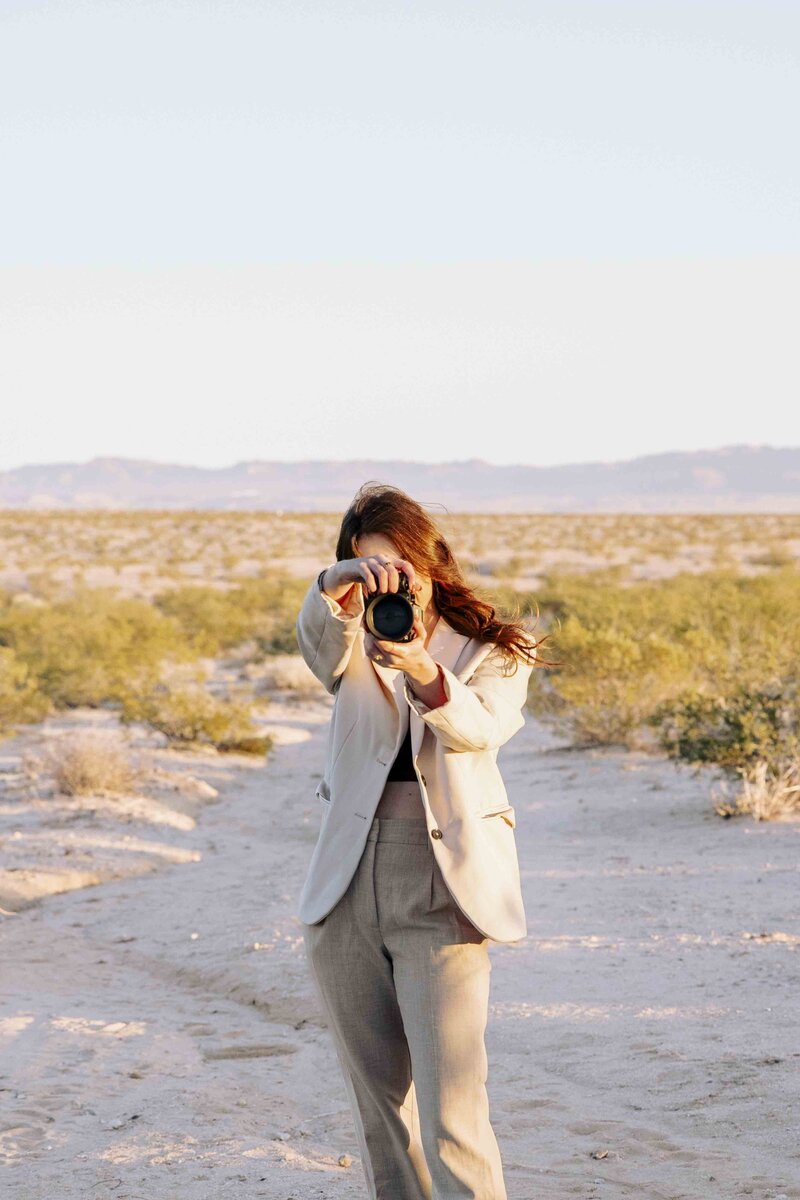 Woman pointing her camera at the viewer with a desert landscape behind her