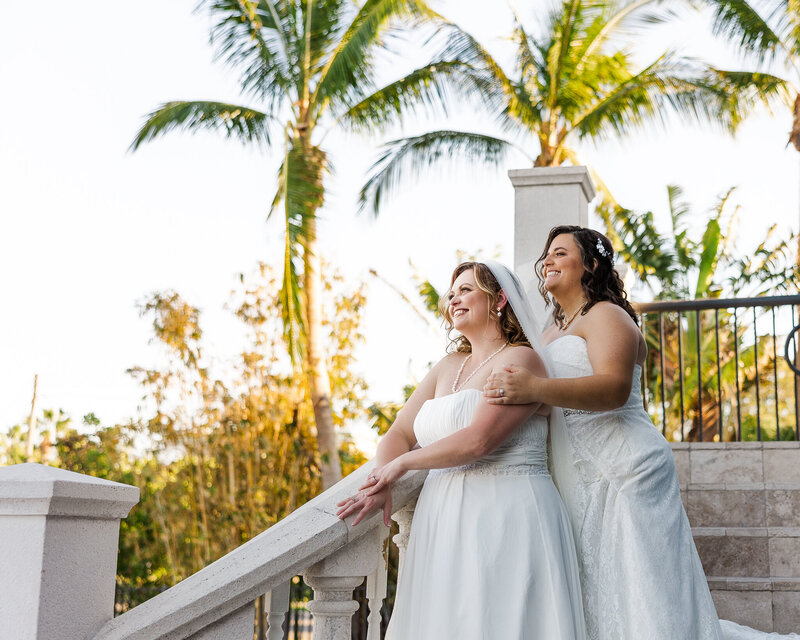 A lesbian couple stand on a white staircase and smile with palm trees in background
