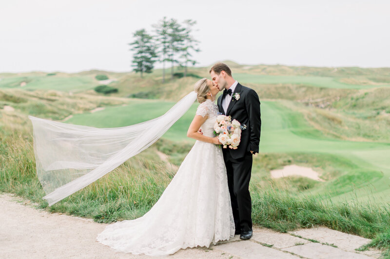 Photo of a bride and groom kissing in front of a beautiful country club golf course. Bride's cathedral veil is blowing in the wind.