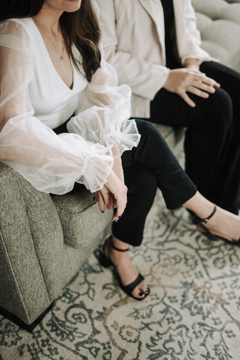 Cropped shot of woman wearing elegant white chiffon top, cropped black pants, and modern black sandal heels sitting on gray couch in Knoxville branding studio Liberty Type