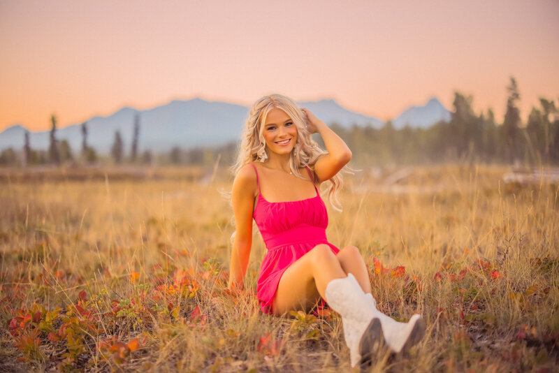 senior girl with long blonde wavy hair wearing a hot pink mini dress sitting in a golden field with blue glacier national park mountains in the background at sunset