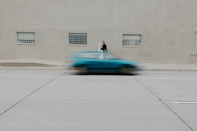 Bride and groom standing against concrete wall outside of Moss Denver kissing with fast moving car in frame